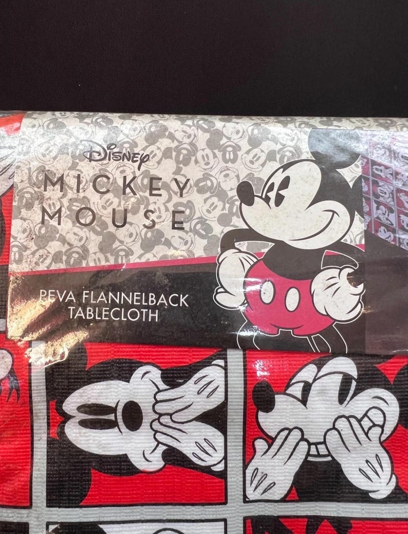 以下が含まれることがあります： ミッキーマウスをテーマにしたテーブルクロス。テーブルクロスは、ミッキーマウスの顔と体の繰り返し模様が赤と黒の背景に描かれています。ディズニーのロゴと「ミッキーマウス」がテーブルクロスの上部に印刷されています。