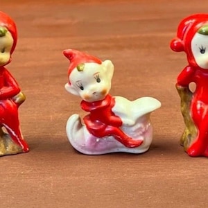 May include: Three ceramic figurines featuring small elf-like characters dressed in bright red suits and pointed hats. Two figures are seated on textured brown tree stumps, while the central figure is positioned inside a small white and pink sleigh.
