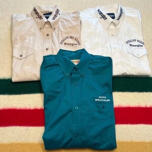 May include: Three long-sleeve button-down shirts. Two are white with Wrangler embroidery on the collars and Puyallup Rodeo Wrangler text on the chests. The third is teal with WSFR Wrangler text embroidered on the chest pocket area.