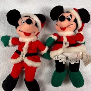 K&ouml;nnte beinhalten: Zwei Pl&uuml;schpuppen von Mickey und Minnie Mouse in Weihnachtsmann-Outfits. Mickey tr&auml;gt eine rote Jacke und Hose mit schwarzem G&uuml;rtel und Stiefeln, gr&uuml;nen Handschuhen und einem Weihnachtsmannhut. Minnie tr&auml;gt ein &auml;hnliches Outfit mit einem wei&szlig;en Rock und gr&uuml;nen Schuhen.