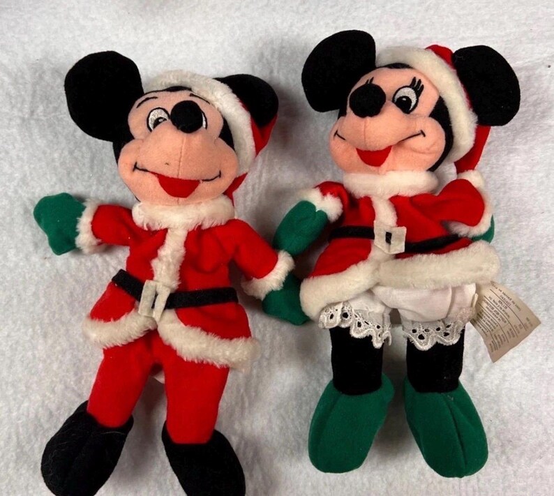 K&ouml;nnte beinhalten: Zwei Pl&uuml;schpuppen von Mickey und Minnie Mouse in Weihnachtsmann-Outfits. Mickey tr&auml;gt eine rote Jacke und Hose mit schwarzem G&uuml;rtel und Stiefeln, gr&uuml;nen Handschuhen und einem Weihnachtsmannhut. Minnie tr&auml;gt ein &auml;hnliches Outfit mit einem wei&szlig;en Rock und gr&uuml;nen Schuhen.