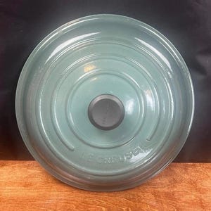 Puede incluir: Una tapa de olla Le Creuset de color verde azulado con un pomo negro. La tapa tiene anillos concéntricos y el nombre de la marca grabado en el lateral. La tapa está sobre una superficie de madera.