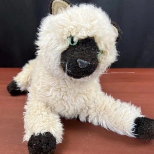 May include: A plush Siamese cat toy with cream-colored fur and dark brown accents on the ears, face, and paws. The stuffed animal features light green eyes, a black nose, and long white whiskers, depicted in a reclining position with one paw extended.