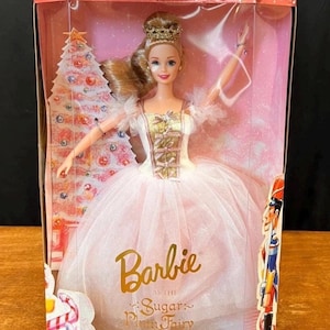 May include: A Barbie doll in a collector edition box, dressed as the Sugar Plum Fairy. The doll wears a white and gold bodice with a layered pink tulle skirt and a gold crown. The packaging features a Christmas tree graphic and text.