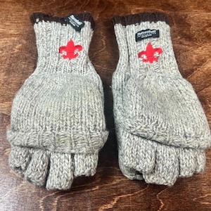 Puede incluir: Un par de mitones convertibles de punto gris con ribete marrón. Cada mitón presenta un emblema de flor de lis rojo y una pequeña etiqueta negra. Las partes superiores de los mitones se pliegan hacia atrás para revelar guantes sin dedos.