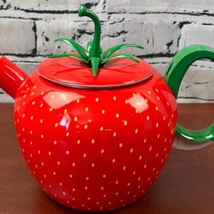 May include: A bright red teapot shaped like a strawberry with small yellow dots across the surface. It features a green handle, a red spout, and a lid topped with a green stem and leaf design that serves as a handle for the lid.