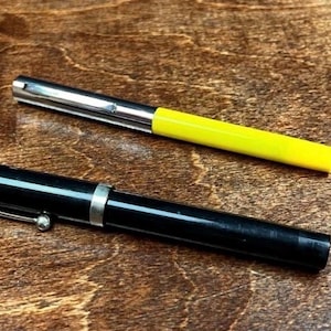 Op de afbeelding: Twee pennen liggen op een houten oppervlak. Eén pen is zwart met een zilveren clip en band. De andere heeft een zilveren punt en een felgele schacht. De pennen staan schuin, wat suggereert dat ze klaar zijn voor gebruik.