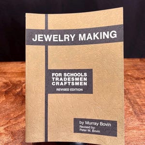 以下が含まれることがあります： 「JEWELRY MAKING」というタイトルの本で、ベージュ色の表紙です。本のタイトルは、黒い横棒の上に白いブロック文字で書かれています。タイトルの下には、「FOR SCHOOLS TRADESMEN CRAFTSMEN REVISED EDITION」というテキストが印刷されています。著者はマレー・ボビンです。