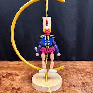 May include: A wooden toy soldier figurine in a blue and pink uniform, suspended from a gold-colored arched frame. The figurine has a white hat with a red feather and is mounted on a wooden base. The toy is a decorative item.