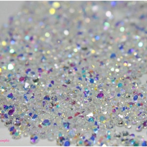 May include: A close-up image of iridescent clear rhinestones. The rhinestones are scattered on a white surface.