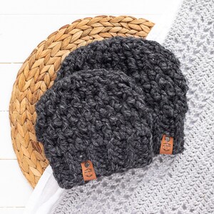 May include: Two gray crocheted hats with brown leather labels. The hats are sitting on a woven straw mat and a white crocheted fabric.