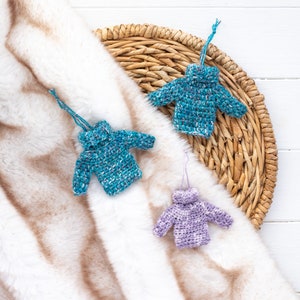 May include: Three crocheted sweater ornaments in blue, teal, and purple hanging from a wicker basket on a white faux fur background.