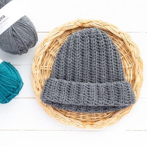 May include: A gray crocheted beanie hat with a ribbed texture sits on a woven wicker basket. The hat has a folded brim and is made with a chunky yarn.