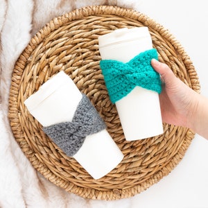 May include: Two white to-go coffee cups with crocheted cozy sleeves. One sleeve is teal blue and the other is gray.