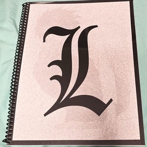 Peut inclure: Un carnet à spirales avec une illustration en noir et blanc de la lettre "L" sur la couverture.