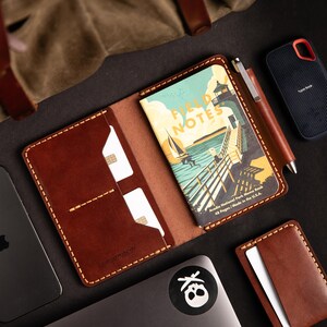 May include: A brown leather wallet with a stitched edge and a pen holder. The wallet has two card slots and a pocket for a small notebook. The notebook is titled "Field Notes" and has a colorful illustration of a lighthouse and a pier. The wallet is on a black surface with a laptop and a small external hard drive.