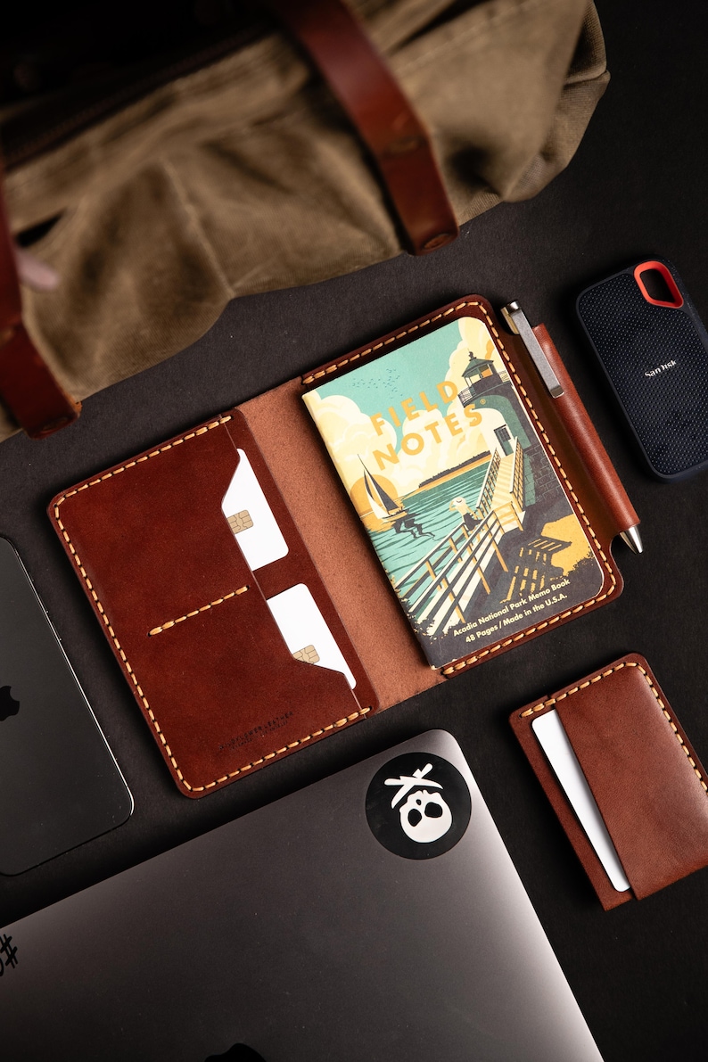 May include: A brown leather wallet with a stitched edge and a pen holder. The wallet has two card slots and a pocket for a small notebook. The notebook is titled "Field Notes" and has a colorful illustration of a lighthouse and a pier. The wallet is on a black surface with a laptop and a small external hard drive.