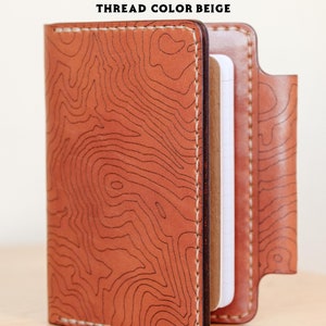 May include: A brown leather notebook cover with a topographic map design and beige stitching. The cover is closed and the notebook is visible inside. "BUCK BROWN + TOPO THREAD COLOR BEIGE" is printed on the cover.