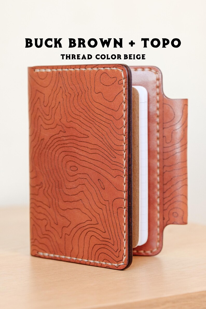 May include: A brown leather notebook cover with a topographic map design and beige stitching. The cover is closed and the notebook is visible inside. "BUCK BROWN + TOPO THREAD COLOR BEIGE" is printed on the cover.