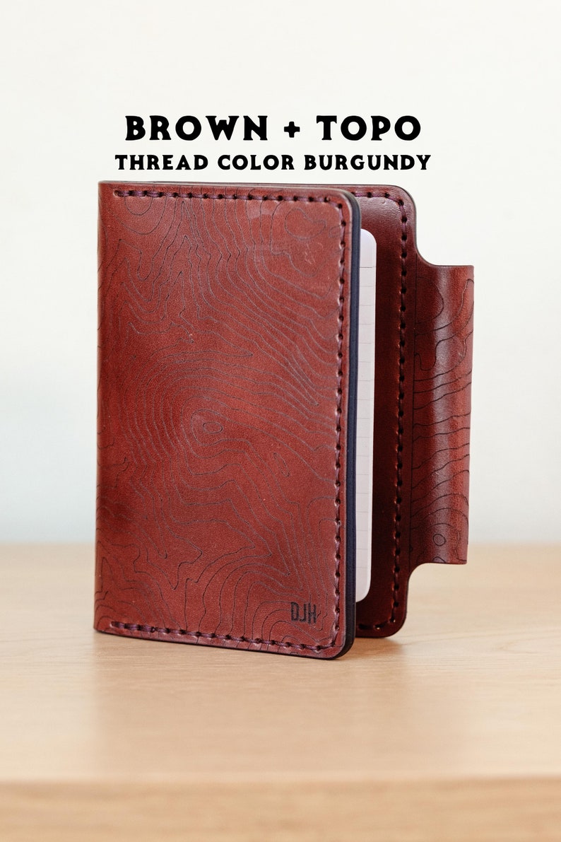 May include: A brown leather notebook cover with a topographic pattern and burgundy stitching. The cover has a pen loop and the initials "DJH" are embossed on the front.