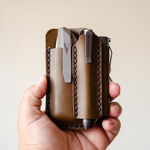 May include: A brown leather pocket organizer with two slots for carrying a pen and a small tool. The organizer has a stitched edge and is held in a person's hand.