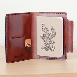 May include: A brown leather notebook cover with a stitched edge and a pocket on the front. The cover is open to reveal a blank notebook with a drawing of an eagle carrying a snake in its talons. The notebook has the text "Carry What Matters" printed on it.