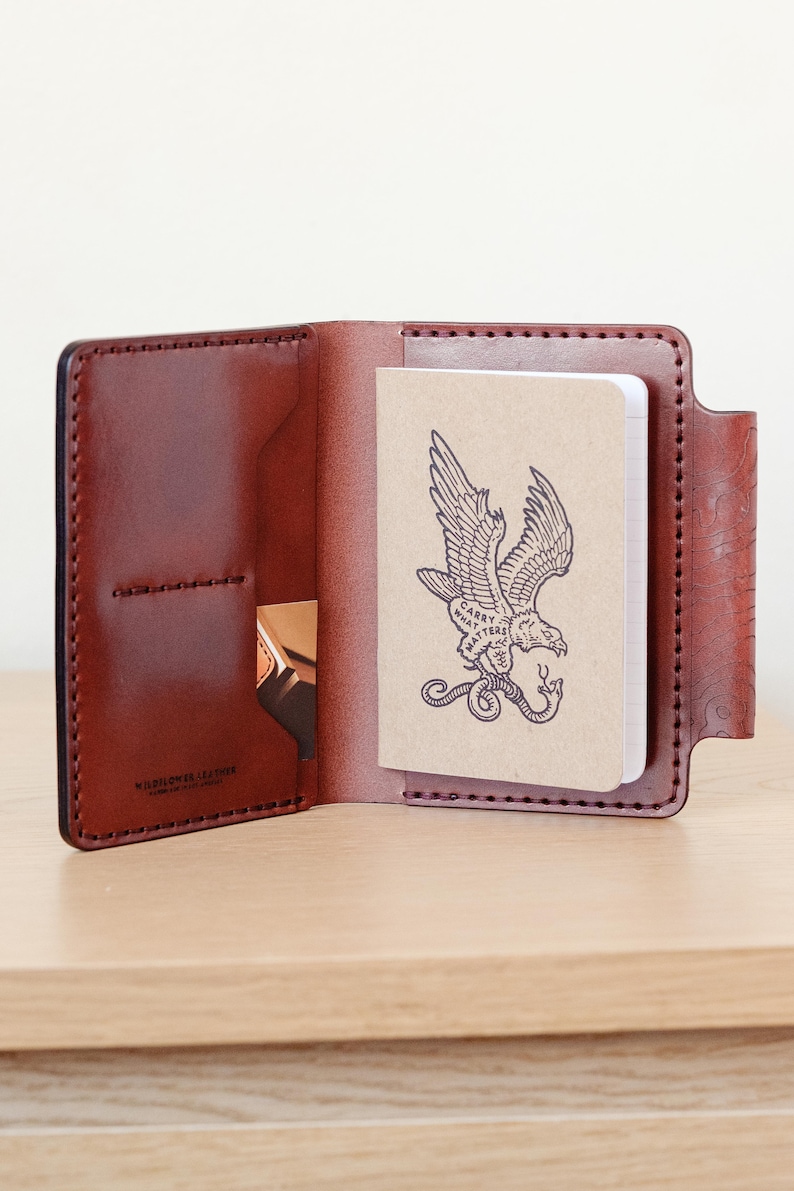 May include: A brown leather notebook cover with a stitched edge and a pocket on the front. The cover is open to reveal a blank notebook with a drawing of an eagle carrying a snake in its talons. The notebook has the text "Carry What Matters" printed on it.