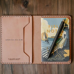 May include: A light brown leather notebook cover with a stitched border. The cover is open to reveal a small notebook with a colorful illustration of a lighthouse and sailboat. A black pen is placed inside the notebook.
