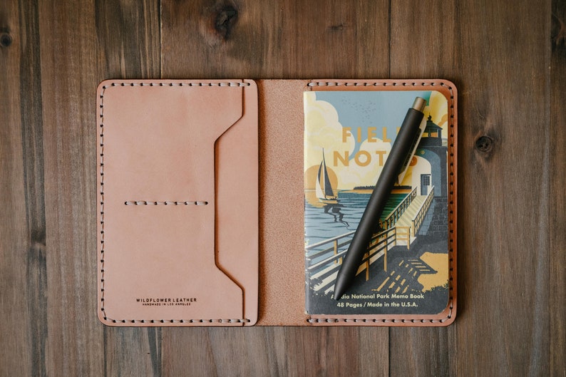 May include: A light brown leather notebook cover with a stitched border. The cover is open to reveal a small notebook with a colorful illustration of a lighthouse and sailboat. A black pen is placed inside the notebook.