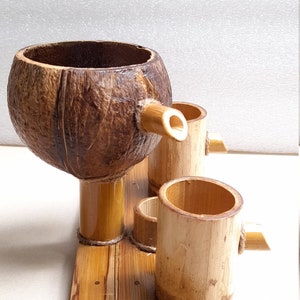 May include: A set of three bamboo cups and a coconut shell cup with a bamboo spout, all sitting on a bamboo tray. The cups are made of natural bamboo and have a light brown color.