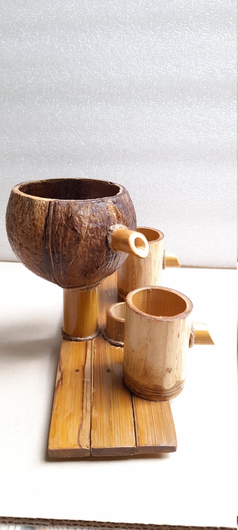 May include: A set of three bamboo cups and a coconut shell cup with a bamboo spout, all sitting on a bamboo tray. The cups are made of natural bamboo and have a light brown color.