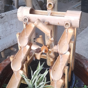 Könnte beinhalten: Ein Bambus-Brunnen mit einem kleinen Wasserrad und mehreren Ebenen mit kaskadierenden Wasser. Der Brunnen ist von grünen Pflanzen umgeben und steht in einem großen braunen Topf.