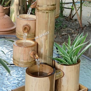 以下が含まれることがあります： 複数の層を持つ竹製の噴水で、水が流れ、植物があります。噴水は自然な木の色で、ガラスの表面に置かれています。「DR SENSEI」の文字が見えます。