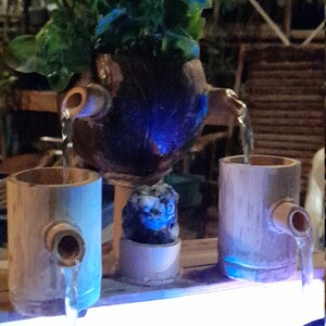 May include: A tabletop water fountain with a blue light. The fountain is made of bamboo and has a coconut shell with a plant growing out of it. The water flows from the coconut shell into two bamboo cups and then into a white bowl.