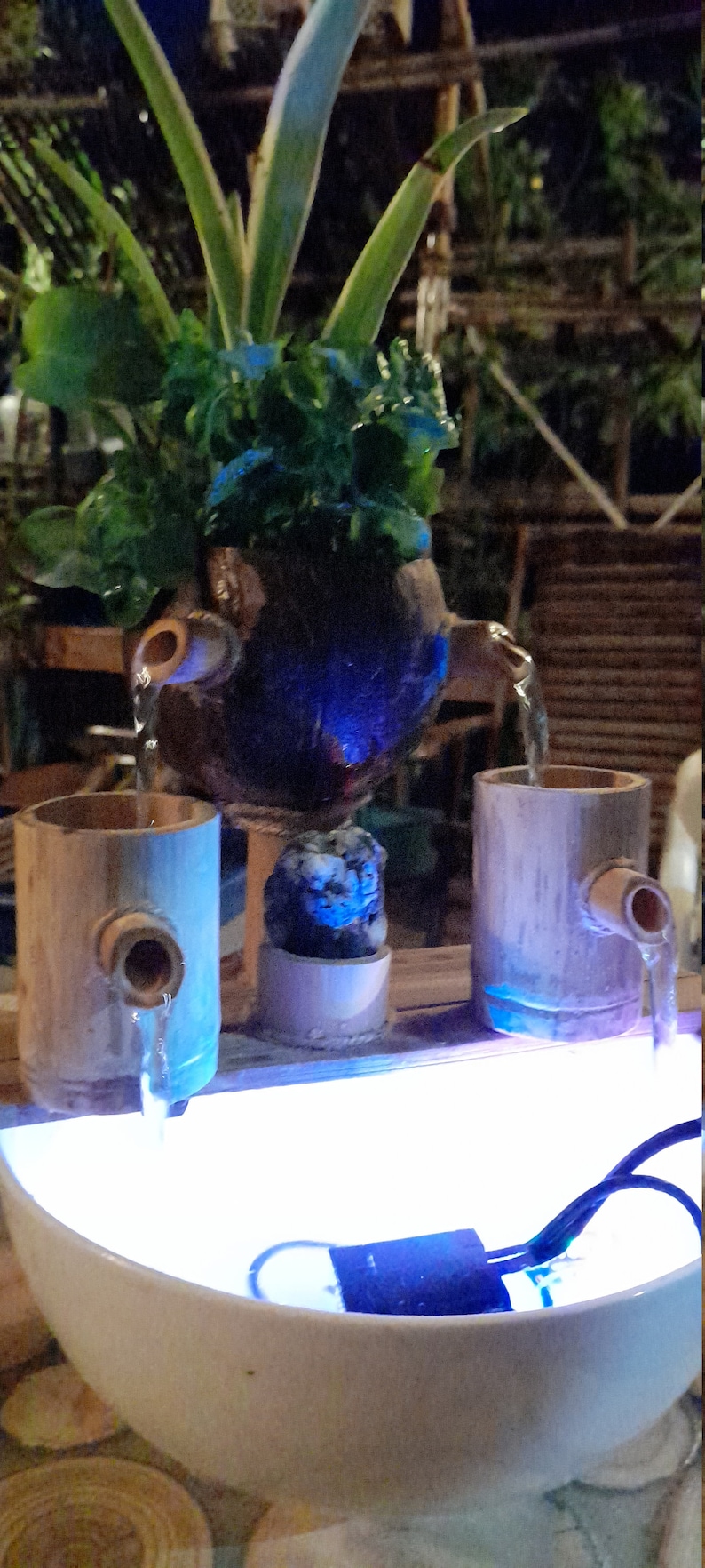 May include: A tabletop water fountain with a blue light. The fountain is made of bamboo and has a coconut shell with a plant growing out of it. The water flows from the coconut shell into two bamboo cups and then into a white bowl.