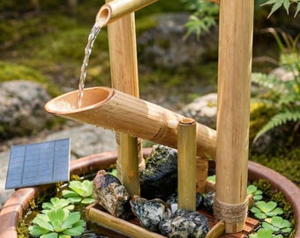 Può includere: Una fontana d'acqua in bambù con una struttura a portale torii e un beccuccio che versa acqua in una vaschetta. La fontana poggia in una ciotola rotonda in terracotta riempita d'acqua, piante acquatiche verdi e pietre scure, con un piccolo pannello solare.