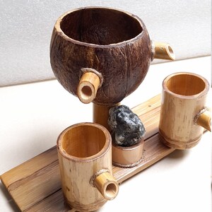 May include: A set of three wooden cups, two made of bamboo and one made from a coconut shell. The cups are connected by a wooden base and have a small spout on each. A small grey stone sits on the base.