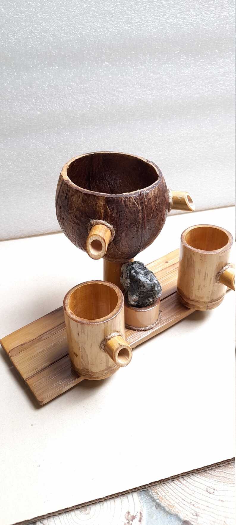 May include: A set of three wooden cups, two made of bamboo and one made from a coconut shell. The cups are connected by a wooden base and have a small spout on each. A small grey stone sits on the base.