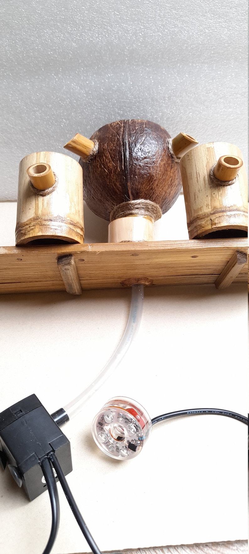 May include: A wooden fountain with a coconut shell as the center piece. The fountain has two bamboo cups on either side of the coconut shell. The fountain is connected to a black pump and a clear tube. The pump is connected to a clear round light with red LEDs.