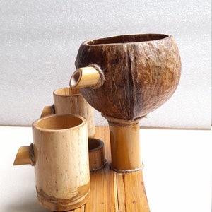 May include: A set of three wooden cups and a coconut shell pitcher, all made of natural bamboo and coconut. The cups and pitcher are arranged on a wooden tray.