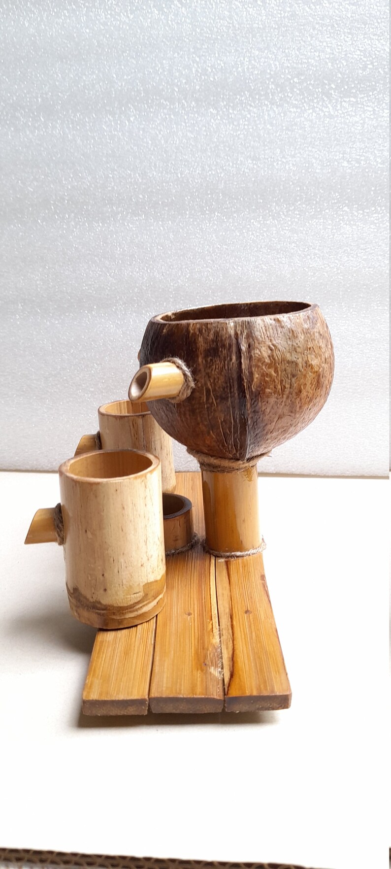 May include: A set of three wooden cups and a coconut shell pitcher, all made of natural bamboo and coconut. The cups and pitcher are arranged on a wooden tray.