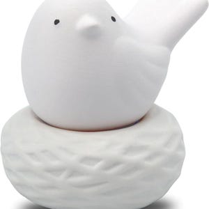 Può includere: Statuetta di uccello in ceramica bianca posata su un nido strutturato. L'uccello ha un corpo arrotondato, un piccolo becco e occhi a puntini neri. Il nido ha un motivo intrecciato. Questo oggetto decorativo è adatto all'arredamento della casa.