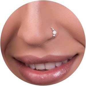 May include: A silver nose ring featuring a central white bead flanked by two smaller silver spheres. The jewelry is positioned on the side of a nostril, showcasing a metallic band that curves along the skin with a polished finish and bead accents.