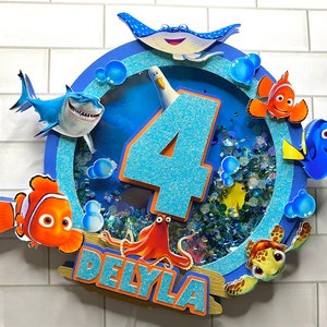 May include: A blue and white cake topper featuring Finding Nemo characters, including Dory, Nemo, and Marlin. The topper also includes a blue number 4 and the name "Delyla".