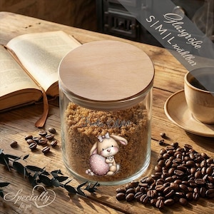 Könnte beinhalten: Ein klares Glas mit einem Holzdeckel, gefüllt mit braunem Zucker. Das Glas hat einen Cartoon-Hasen, der ein rosa Ei hält. Der Text "Frohe Ostern" befindet sich auf dem Glas. Kaffeebohnen und eine Tasse Kaffee stehen auf dem Holztisch.