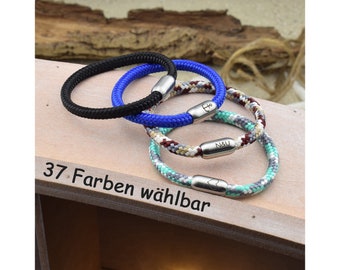 Könnte beinhalten: Drei geflochtene Seilarmbänder mit silberfarbenen magnetischen Metallverschlüssen. Ein Armband ist einfarbig schwarz, eines einfarbig blau mit Ankergravur und eines ist mehrfarbig geflochten mit N&U- und Herzgravuren auf den Verschlüssen.
