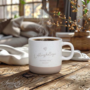 Könnte beinhalten: Ein weißer Keramikbecher mit Sprenkeloptik und einem hellbraunen Boden. Der Becher zeigt ein kleines Herz-Symbol über dem Text "Lieblingskollegin" und "DANKE FÜR ALLES!" in dunkelgrauer Schreib- und Blockschrift. Dampf steigt aus dem dunklen Kaffee auf.