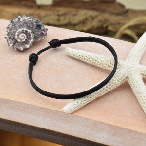 May include: A black braided cord bracelet with a simple knot closure. The bracelet is displayed on a wooden surface with a seashell and a starfish.