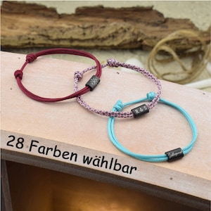 Könnte beinhalten: Drei verstellbare Kordelarmbänder in Bordeaux, einem rosa-grau gesprenkelten Muster und hellem Türkis. Jedes Armband hat eine kleine, rechteckige schwarze Metallperle mit Gravuren, darunter ein Kompass, die Initialen R & S und ein Schriftzug.