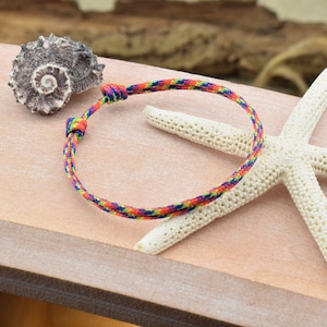 May include: A colorful braided friendship bracelet with a knot closure. The bracelet is made of a thin, multi-colored cord and is shown on a wooden surface with a seashell and a starfish.